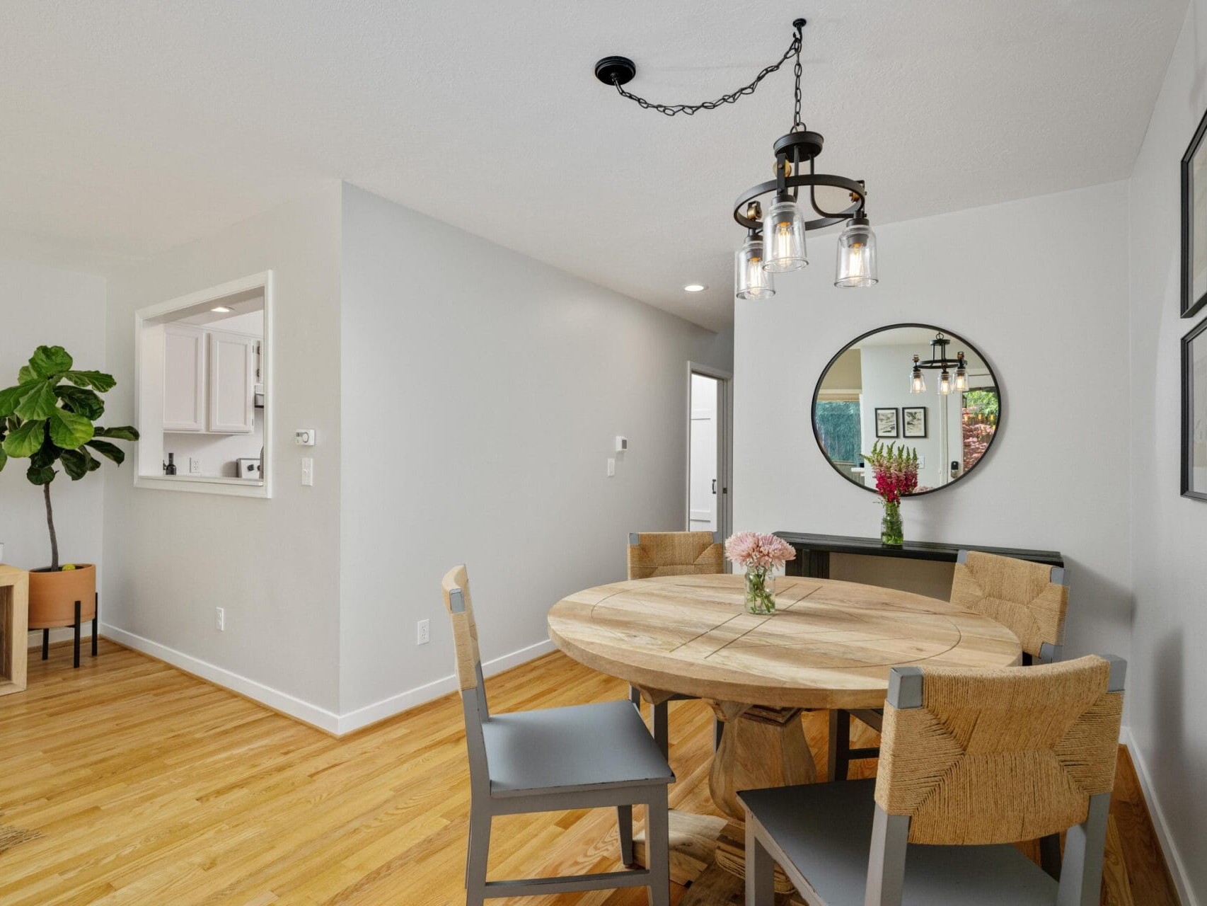 A cozy dining area with a round wooden table, four chairs, a modern chandelier, a mirror on the wall, and a vase of flowers, set on light wood flooring with a view into a white kitchen.