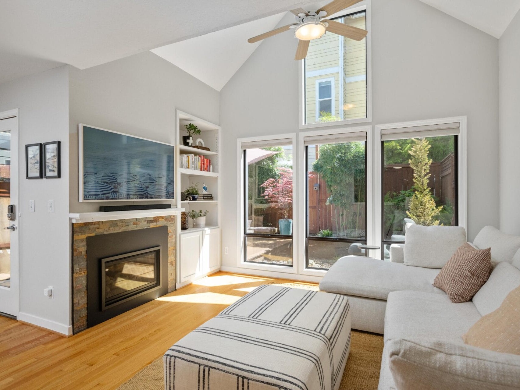 A bright living room with large windows, a beige sectional sofa, a striped ottoman, a fireplace with a TV above it, built-in shelves, and a view of a sunny backyard with greenery.