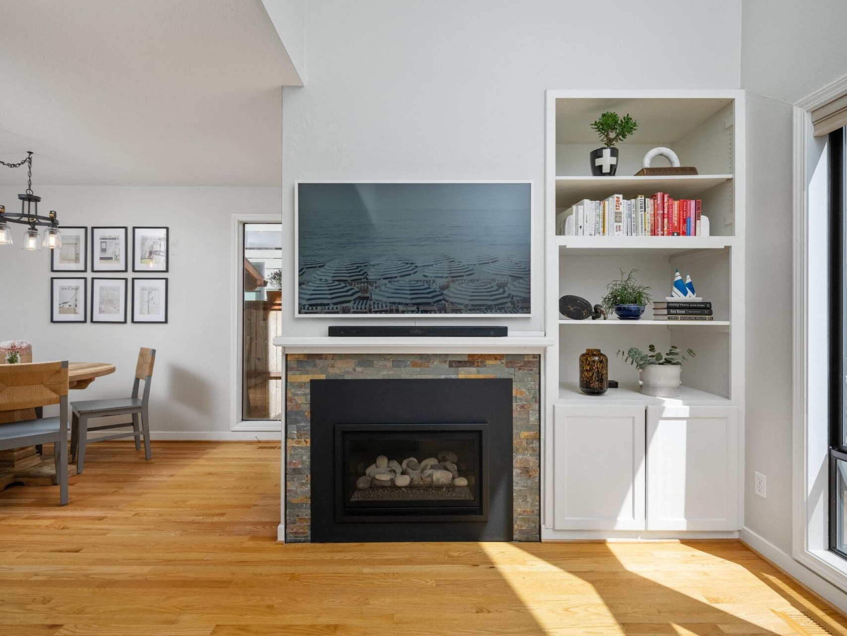 Bright living room with a fireplace beneath a mounted TV, built-in shelves with books and decor on the right, wooden floors, and a dining area with a round table and framed art on the wall to the left.
