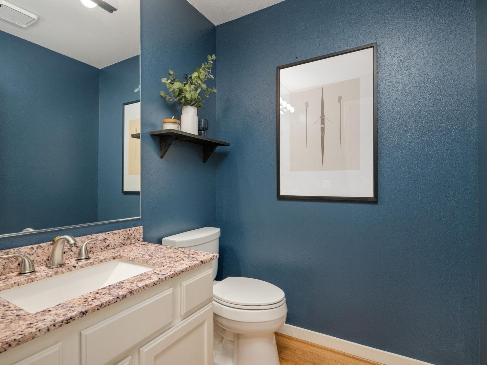 A modern bathroom with dark blue walls, a white toilet, white vanity with a pink granite countertop, large mirror, abstract wall art, and a small black shelf holding decor and greenery.