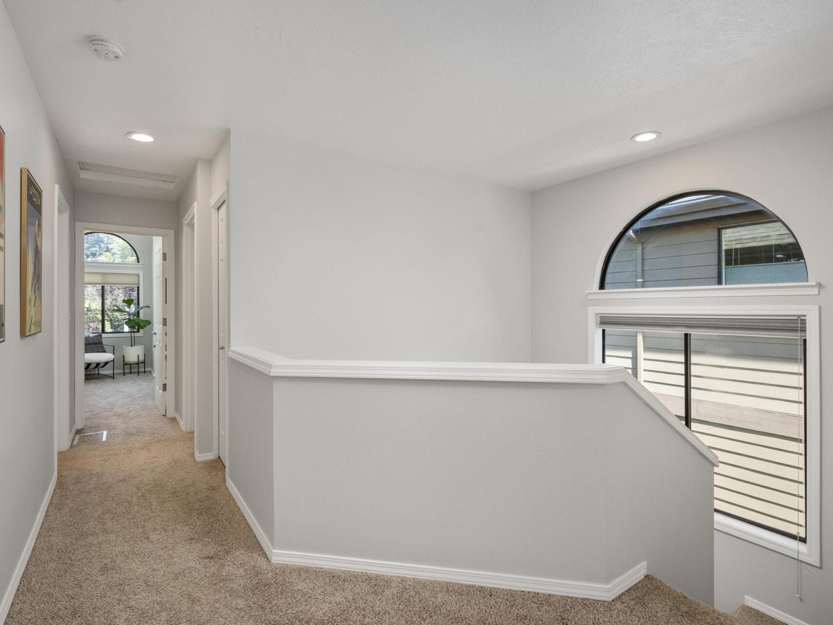 Bright upstairs hallway with white walls, beige carpet, recessed lighting, and a large arched window. Framed art hangs on the wall, and a view into another room with a plant is visible at the hallway’s end.