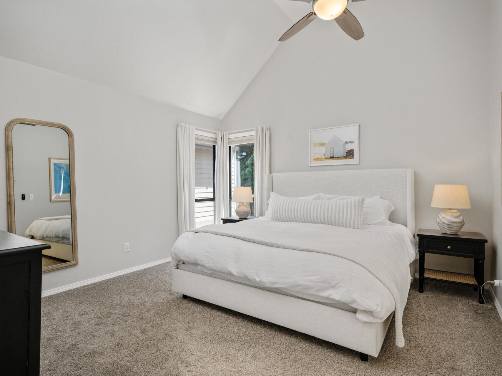 A bright, neutral-toned bedroom with a large upholstered bed, two black nightstands with lamps, a dresser, a tall mirror, and a window with white curtains letting in natural light.