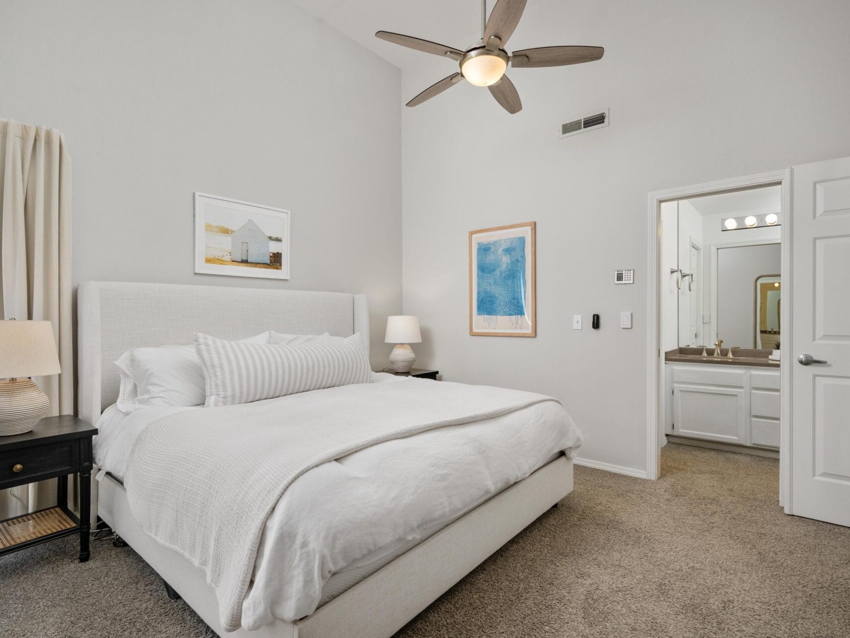 A modern bedroom with a large white bed, beige carpet, two black nightstands with lamps, wall art, and an attached bathroom. A ceiling fan and natural light from a window enhance the bright, airy space.