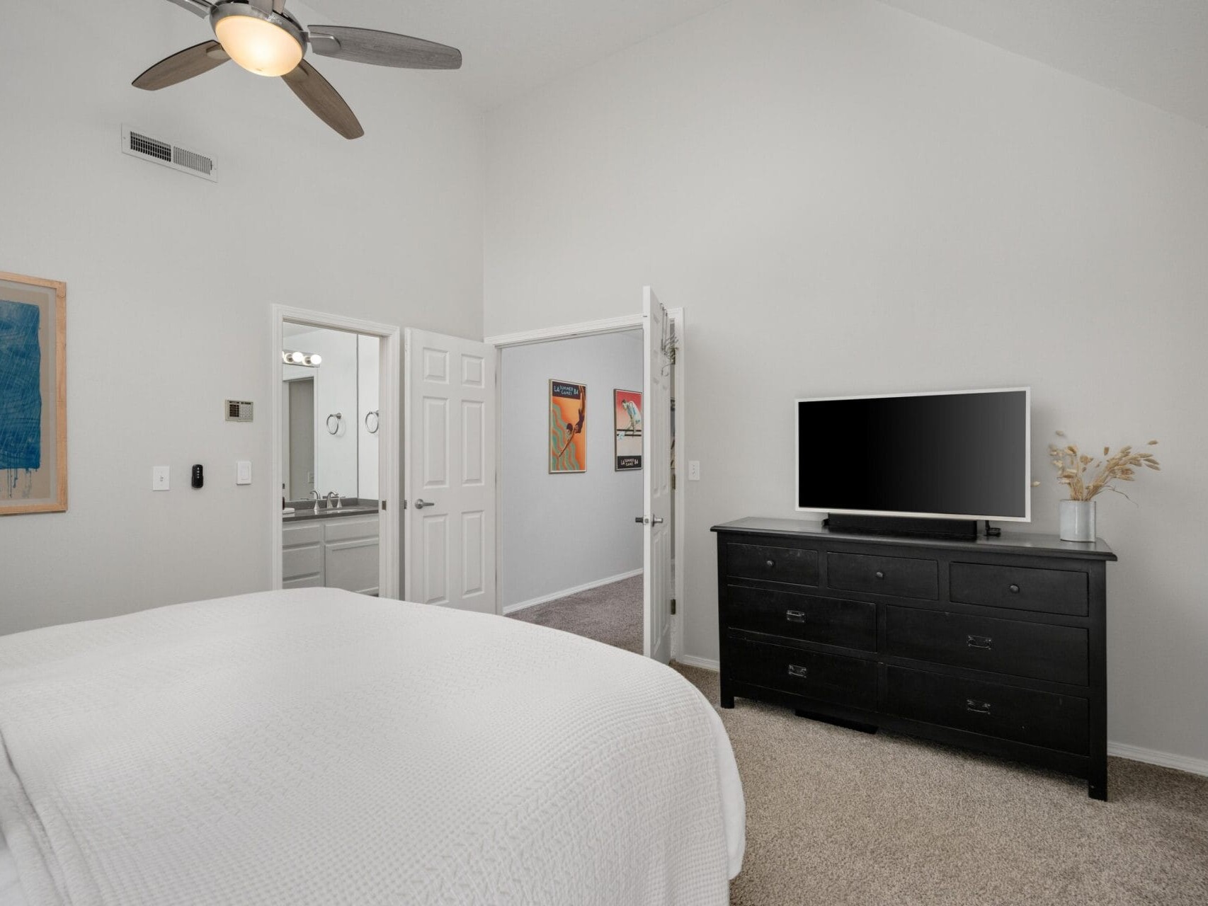 A modern bedroom with a neatly made bed, white bedding, black dresser with a TV, a ceiling fan, and an open door showing a bathroom and hallway with artwork on the walls.