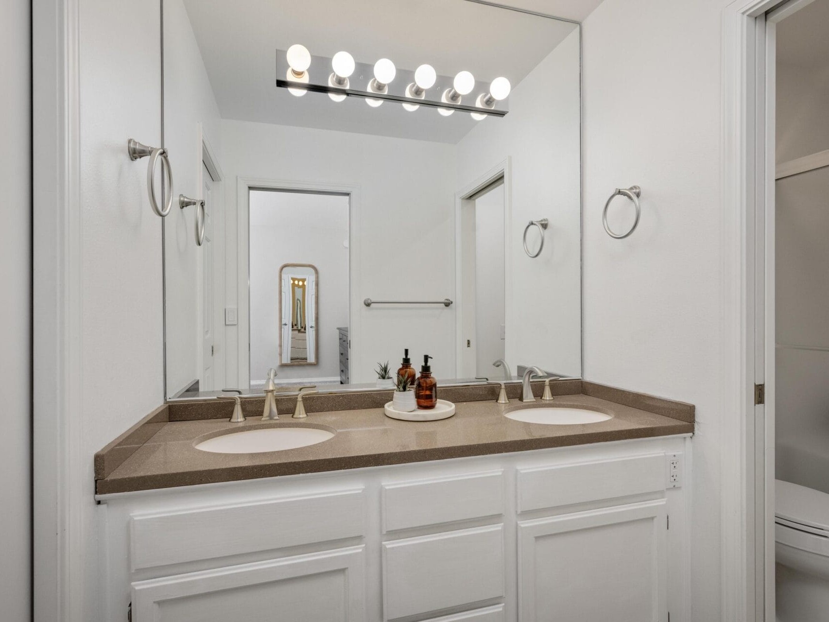 A modern bathroom with a double sink vanity, beige countertop, round mirrors, wall-mounted light fixture, and decorative soap dispensers on a tray. White cabinetry and neutral walls provide a clean look.