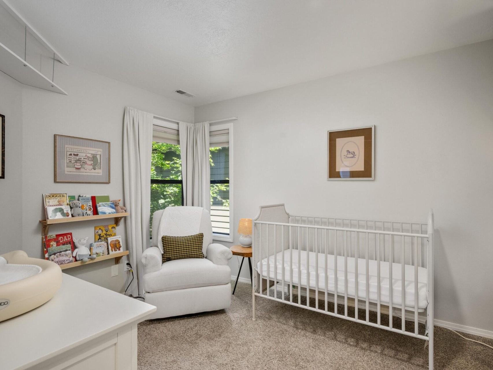 Bright nursery room with white walls, a crib, a cushioned rocking chair with a pillow, bookshelves with children’s books, framed art on the walls, and large windows with light curtains letting in natural light.