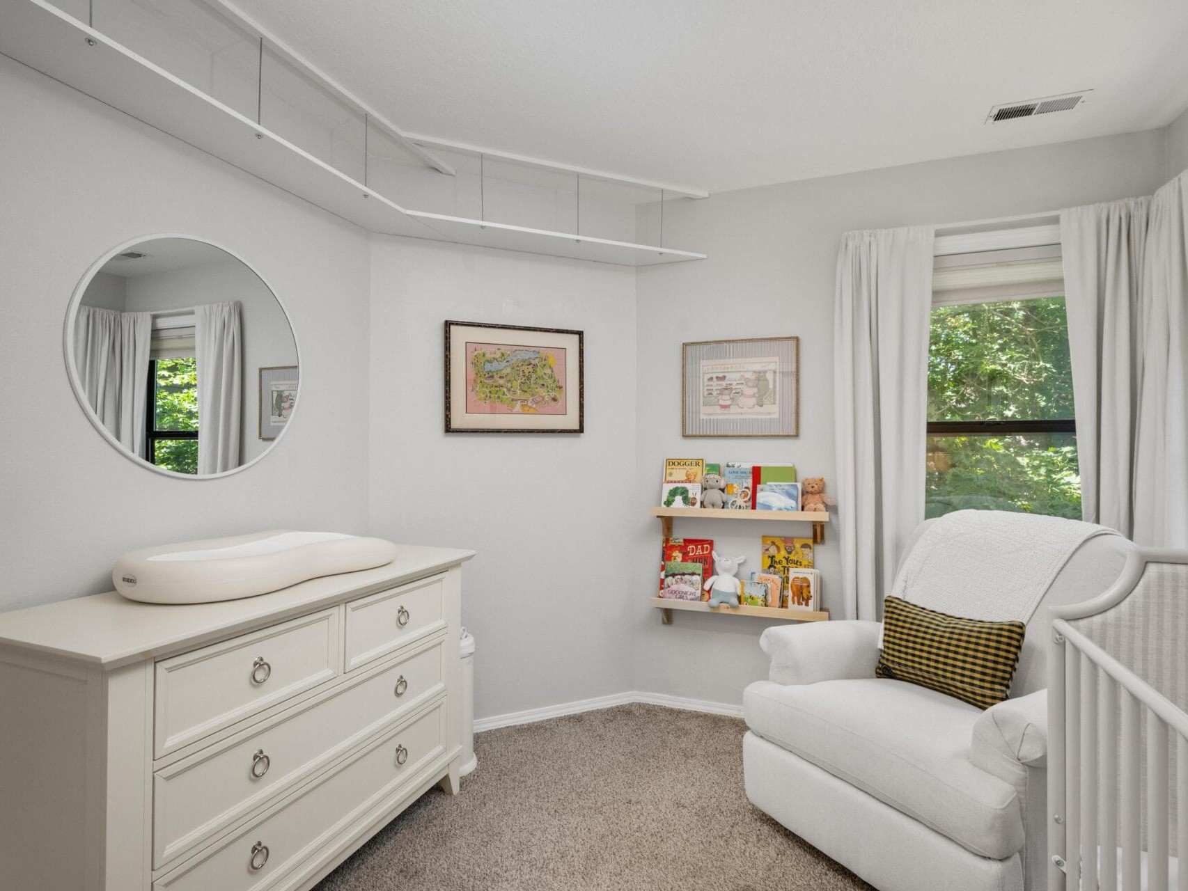 A cozy, light-filled nursery with a white dresser and changing pad, a round mirror, bookshelf with children’s books, a white crib, and a cushioned armchair with a yellow pillow next to two windows with white curtains.