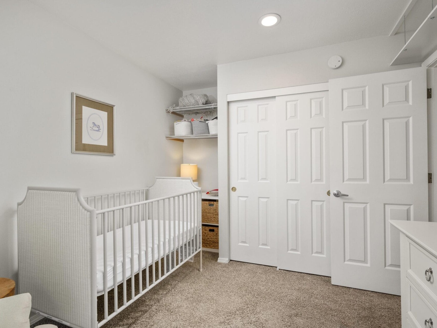 A minimalist nursery with a white crib, beige carpet, a dresser, closet with white sliding doors, shelves holding baskets and blankets, a small lamp, and framed art on the wall. The room is softly lit and neatly organized.