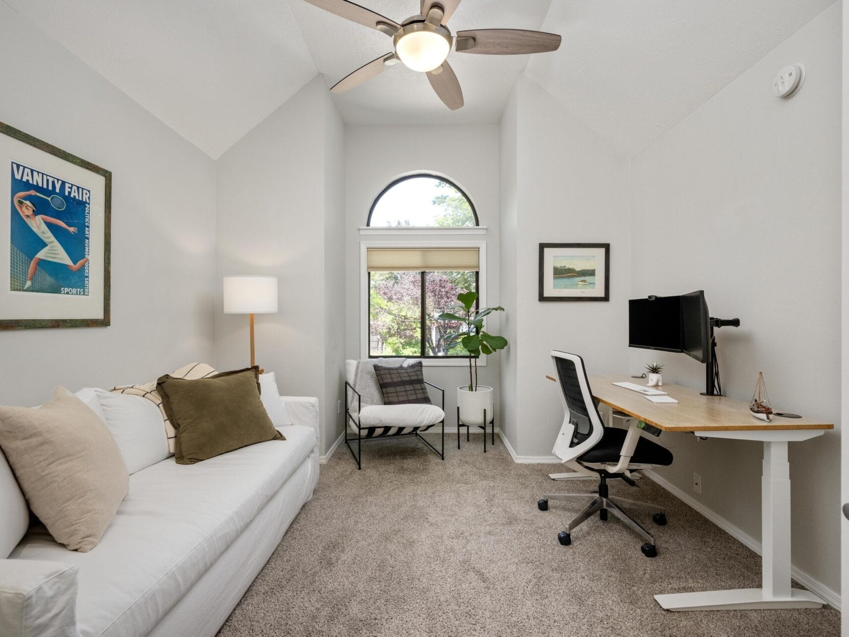 A modern home office with a white couch, armchair, and desk facing a computer. A plant and art decorate the space, and a large arched window lets in natural light. The room has light walls and carpeted floor.