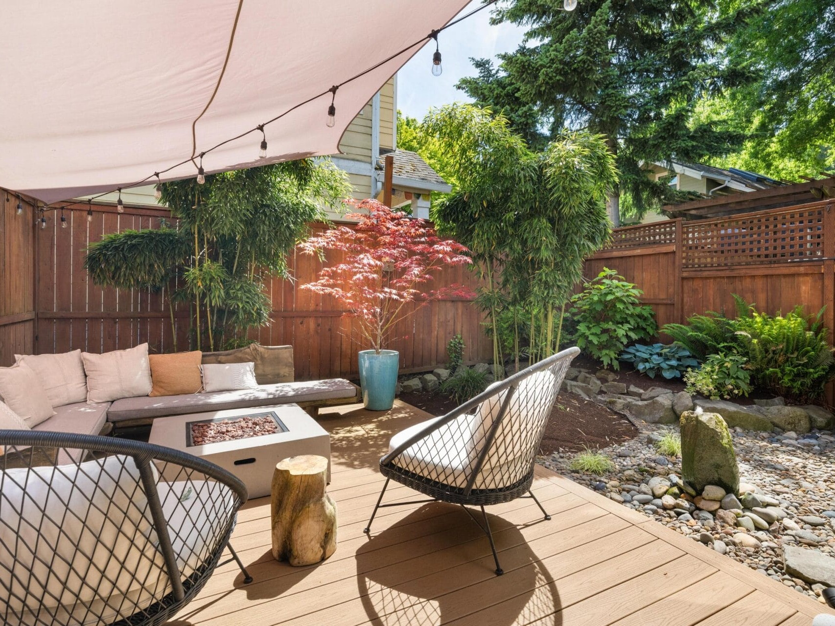 A cozy backyard patio with a wooden deck, modern chairs, a fire pit table, cushioned seating, potted plants, string lights, and a landscaped garden with rocks and trees surrounded by a wooden fence.