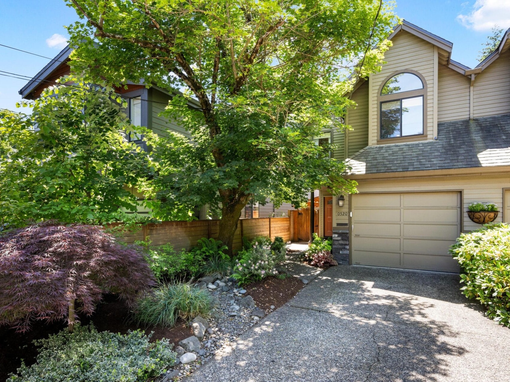 A two-story townhouse with beige siding, a double garage, arched windows, lush landscaping, and a large tree partially shading the front driveway on a sunny day.