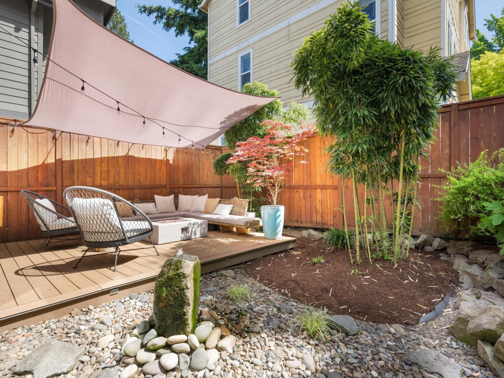 A cozy backyard patio with wicker chairs, a small sofa, string lights, and a beige shade sail. The area features a wooden deck, potted plants, bamboo, a Japanese maple, a rock garden, and a wooden fence.