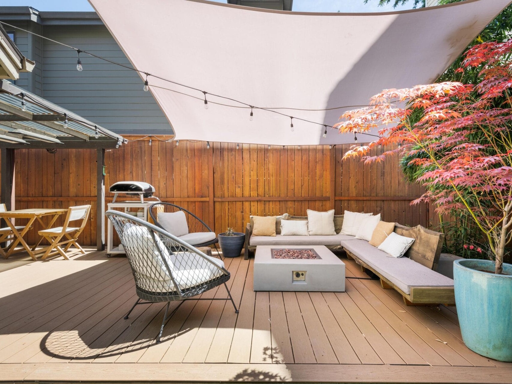 Modern outdoor patio with a wooden deck, lounge seating, a fire pit, string lights, a dining table, a grill, and a large potted plant under a white shade sail.