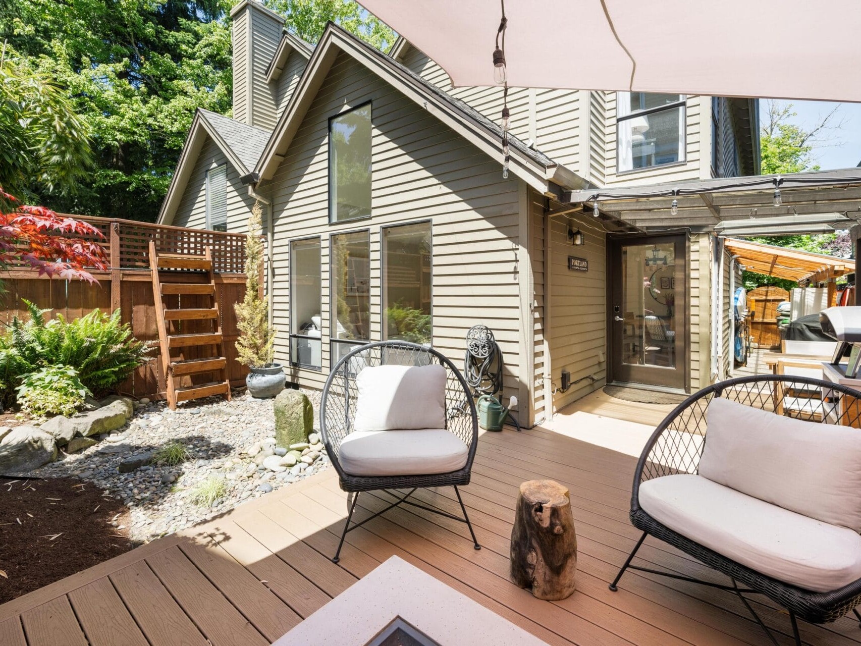 Modern patio with two cushioned chairs, a small wooden table, and landscaped garden. The beige house has tall windows, wood siding, and a stairway leading to a fenced area. Trees and plants surround the outdoor space.