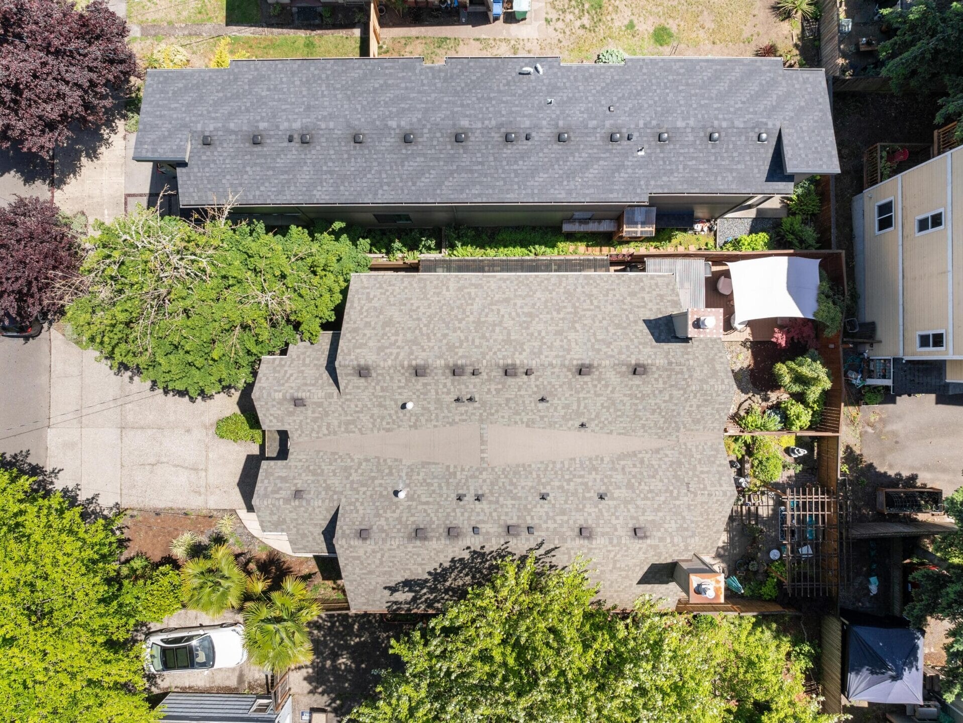 Aerial view of two adjacent rectangular buildings with gray roofs, surrounded by trees and greenery. A driveway with a white car is visible at the bottom left, and a white canopy covers a patio area on the right.