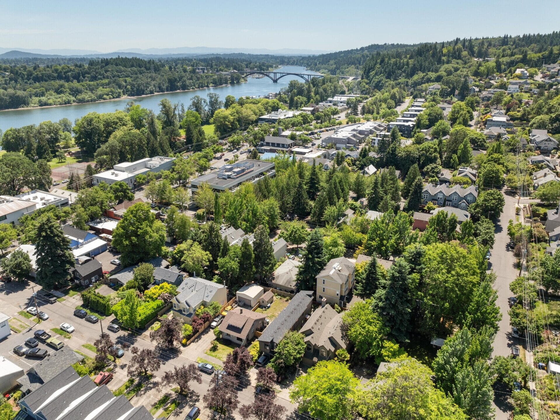 Aerial view of a lush green suburban neighborhood with houses, trees, a river, and a bridge in the distance. Rolling hills and a clear sky are visible in the background.