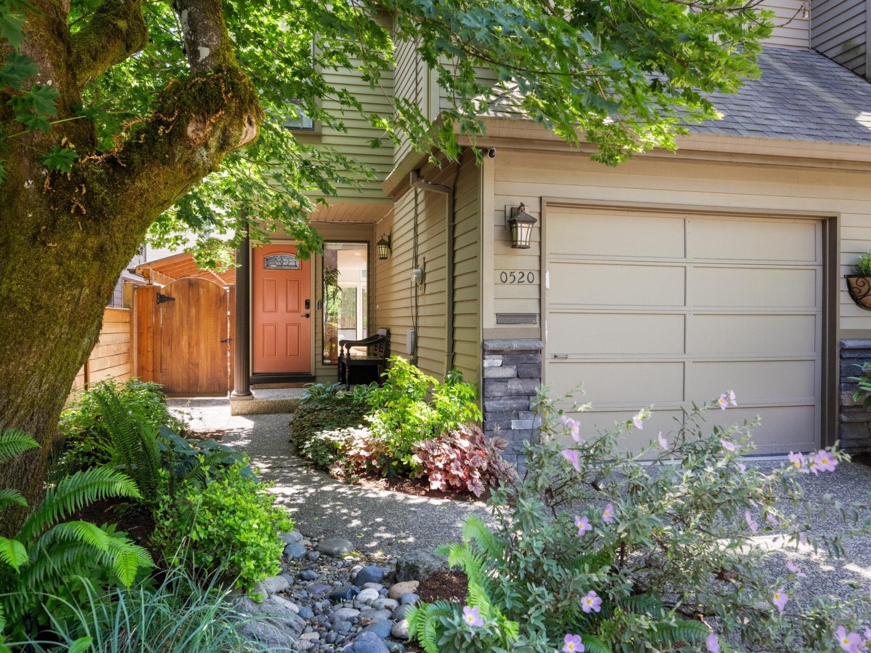 A suburban house with a beige garage, a bright coral front door, stone accents, lush green plants, and trees along a pebbled walkway leading to the entrance.