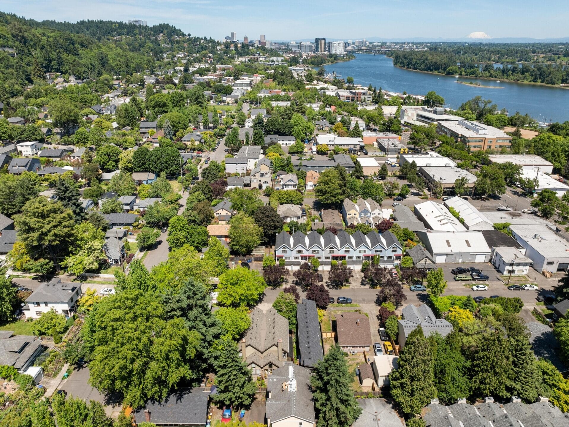 Aerial view of a green suburban neighborhood with houses, trees, and some commercial buildings, beside a river. City buildings and distant hills are visible under a clear blue sky.