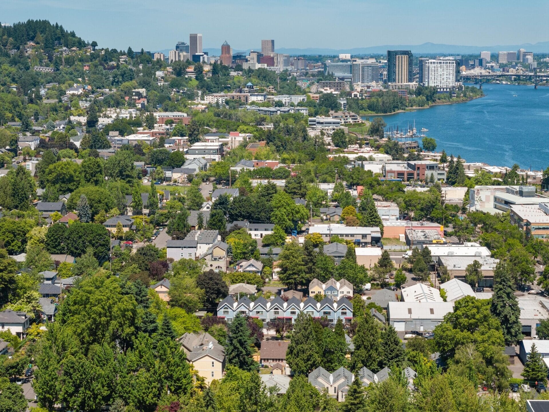 Aerial view of a city with a mix of houses, trees, and buildings near a river, with a hilly, forested area on the left and a downtown skyline in the background under a clear sky.