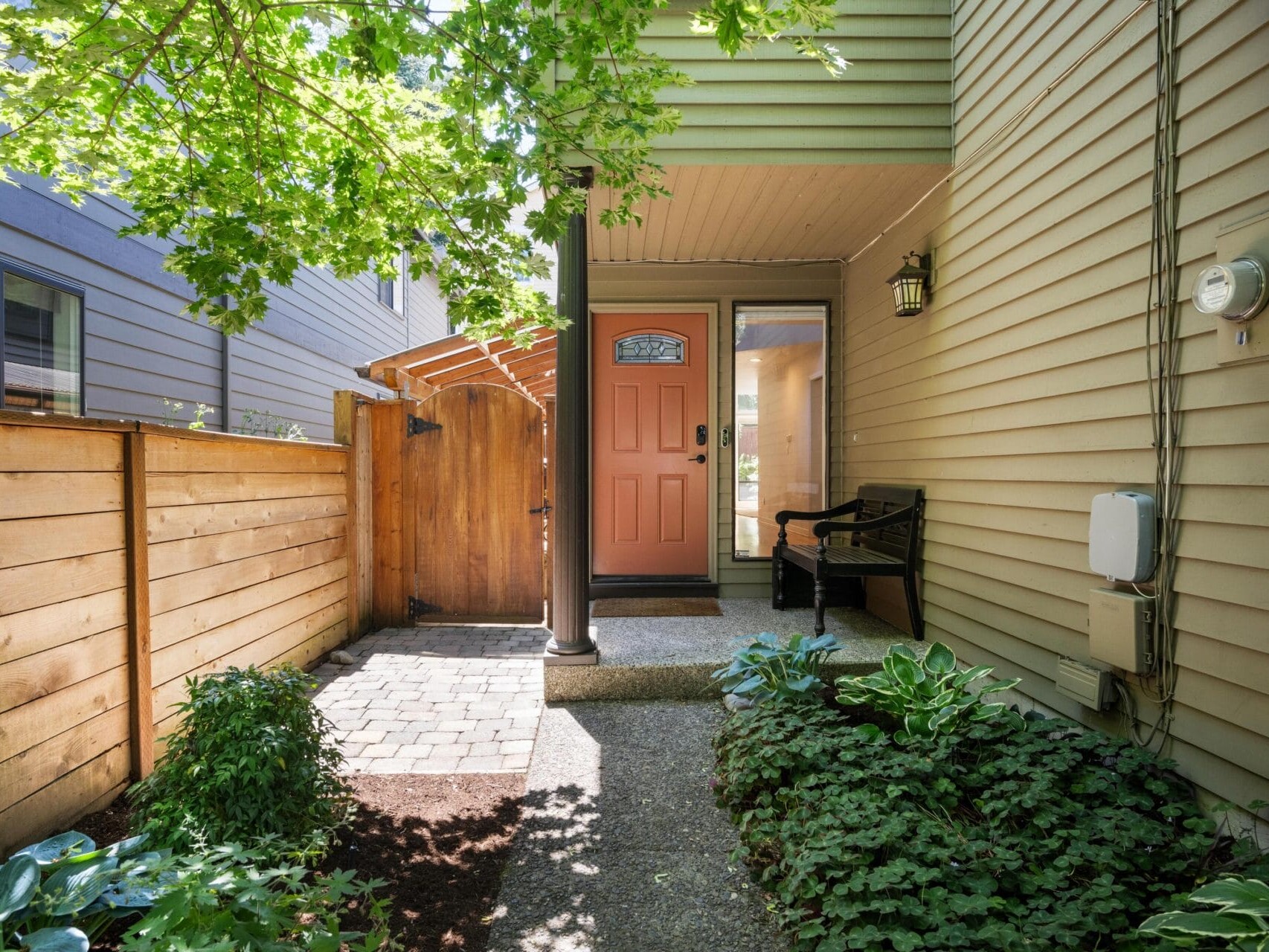 Shaded front entryway of a house with a coral-colored door, a wooden fence and gate, green plants along the path, a black bench, and tan siding. Sunlight filters through tree leaves overhead.