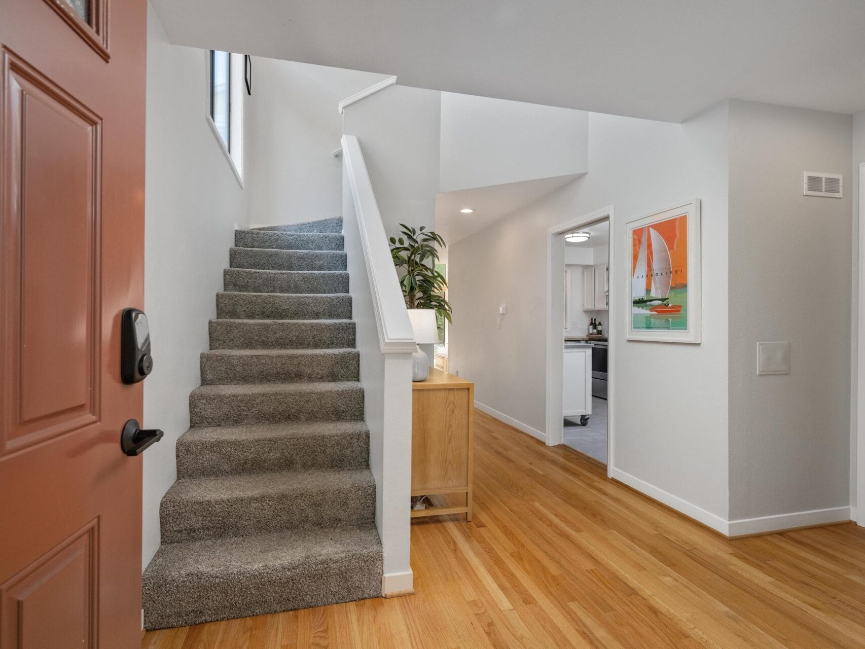 A view from the front door showing a carpeted staircase on the left, light wood flooring, a wooden cabinet with a plant, white walls, a framed sailboat painting, and a glimpse into a kitchen with white cabinets.