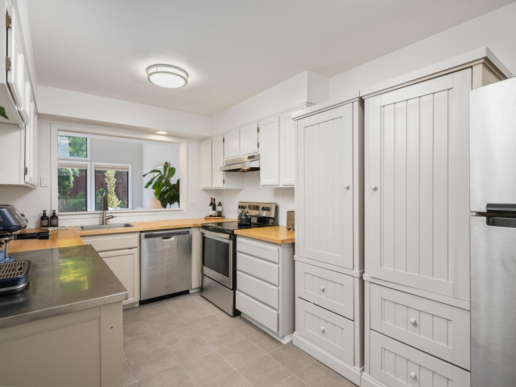 Bright, modern kitchen with white cabinets, stainless steel appliances, light wood countertops, a window above the sink, and potted plants adding greenery to the space.
