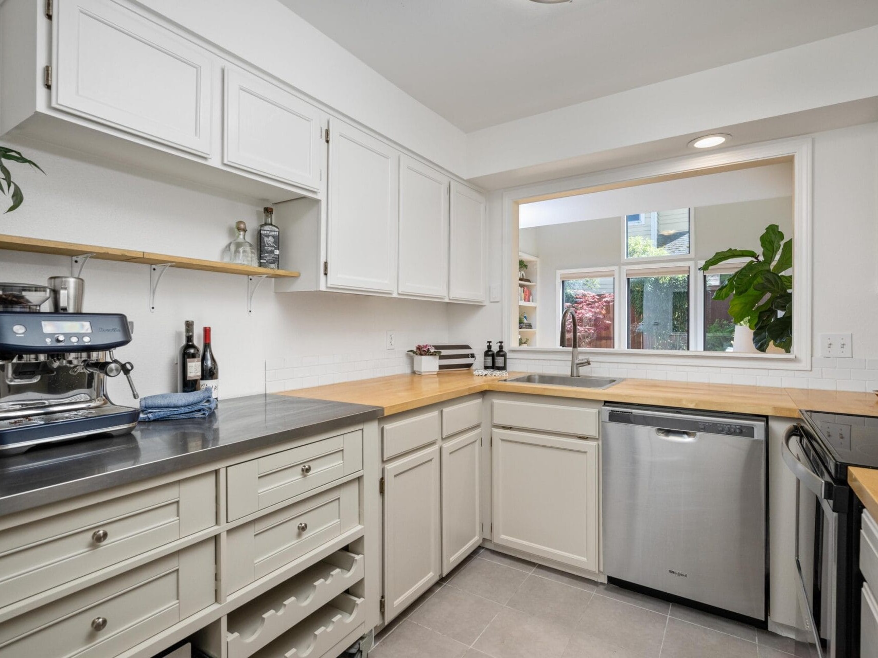Modern kitchen with white cabinets, wood countertops, stainless steel appliances, a coffee machine, open shelving, and a large window above the sink showing greenery outside.