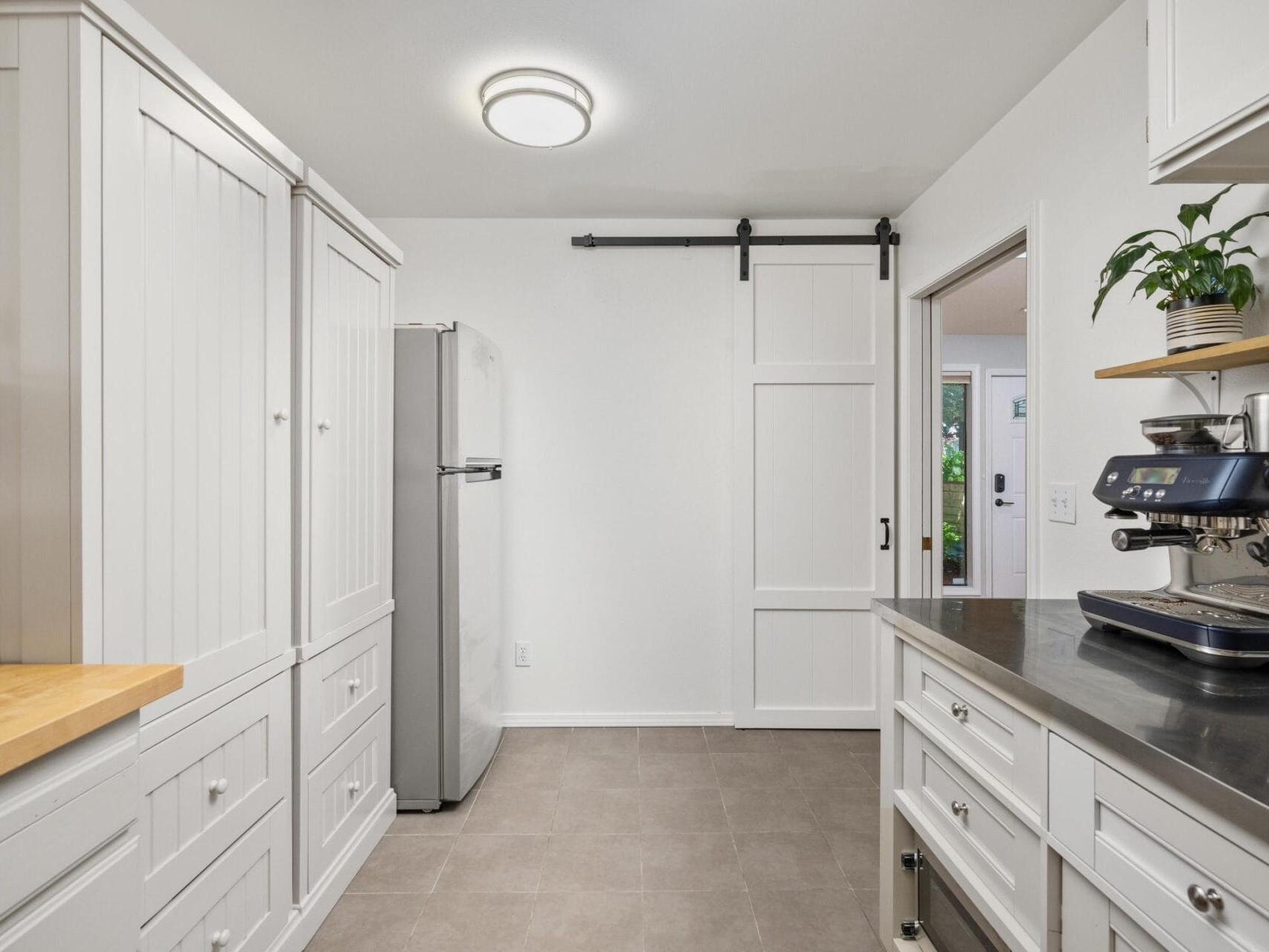 Modern white kitchen with a gray tile floor, stainless steel appliances, a white sliding barn door, a coffee machine on the counter, potted plant on a shelf, and a view into another room.