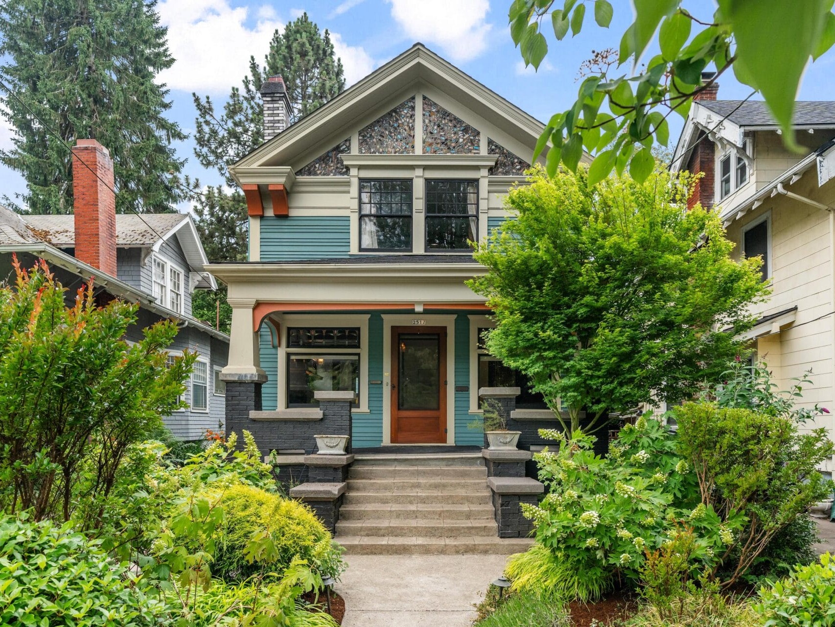 A two-story house with stained glass windows, a front porch, and steps leading to a wooden door, surrounded by lush green bushes and trees on a sunny day.