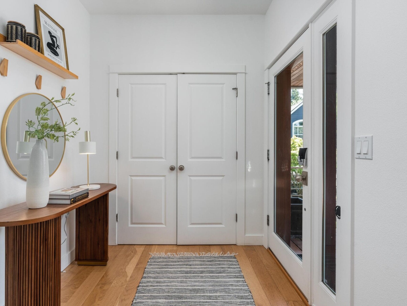A bright entryway with double white doors, a wooden console table with a round mirror above it, a white vase with green branches, a striped rug on a wood floor, and glass doors leading outside.