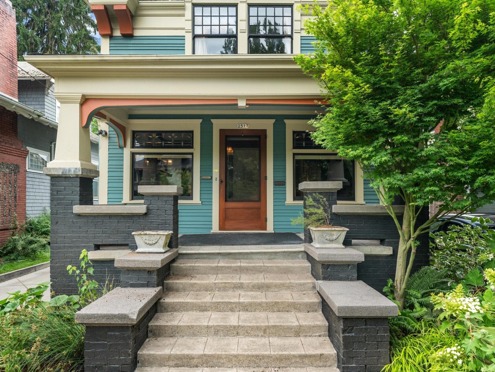 A two-story Craftsman-style house with green and cream siding, wide front steps, a wooden door, black brick columns, and lush landscaping, including a leafy tree near the entryway.