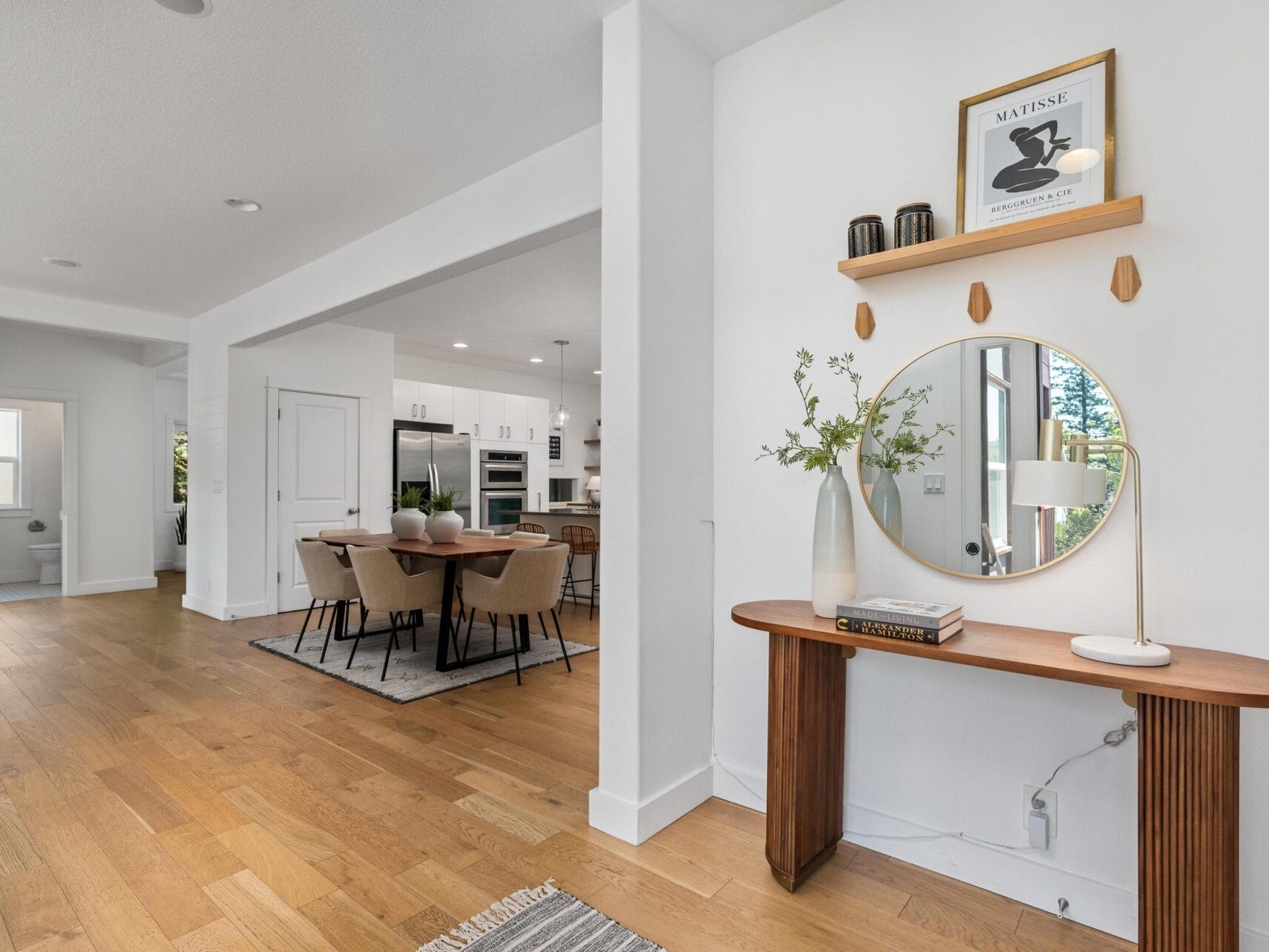 A modern entryway with a round mirror above a wooden console table, decorated with books, a vase with greenery, and a lamp. In the background, a dining area and kitchen with white cabinets are visible.