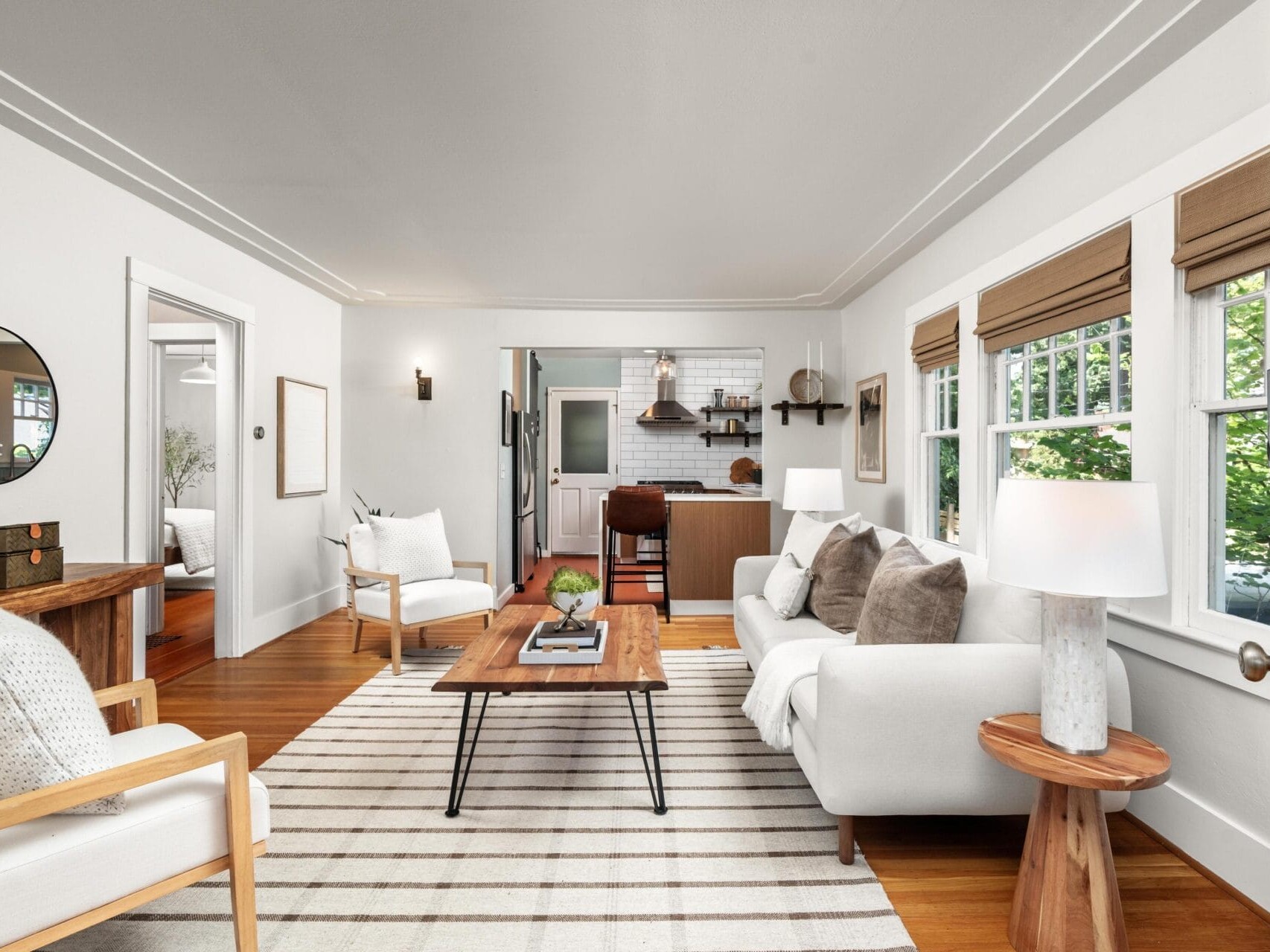 A bright, modern living room with a white sofa, armchairs, a striped rug, and wood accents. Large windows with Roman shades let in natural light. The open layout shows a kitchen with bar stools in the background.