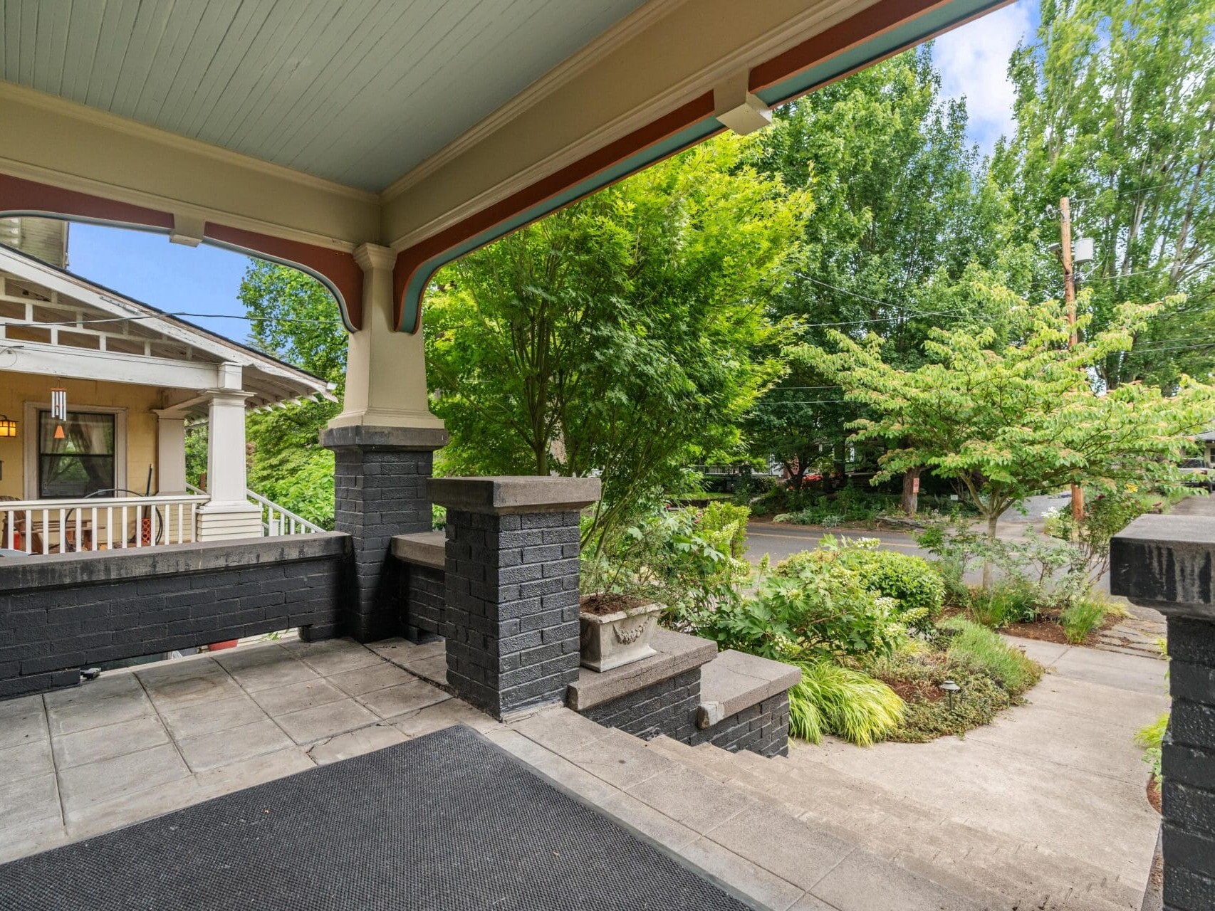 View from a covered front porch with black brick columns, looking out onto a garden with green trees and shrubs, neighboring house, and a sidewalk leading to a quiet street.