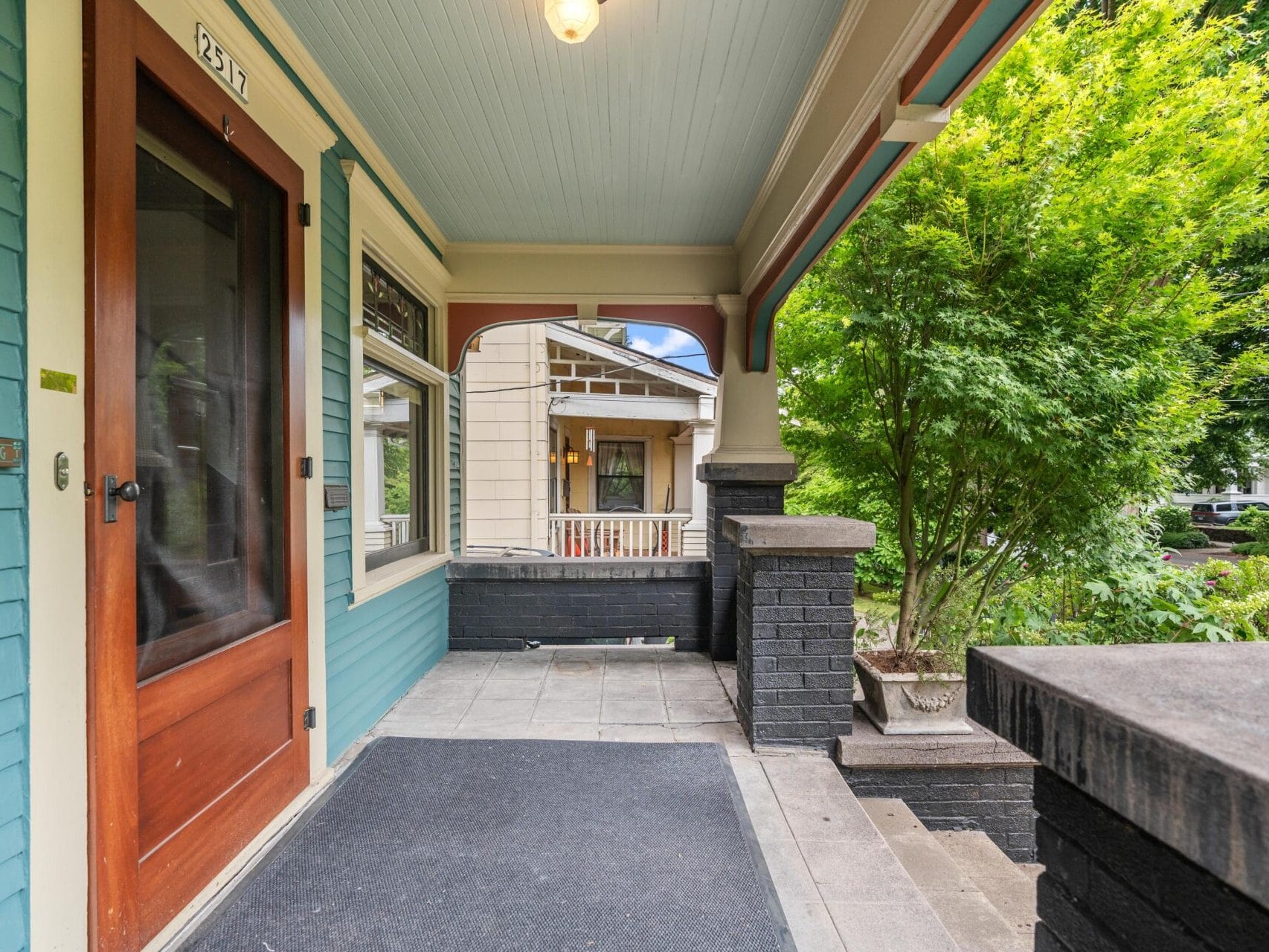 Front porch of a house with teal siding, wooden door, black brick columns, gray floor tiles, a doormat, and green trees nearby; another house is visible across the street.