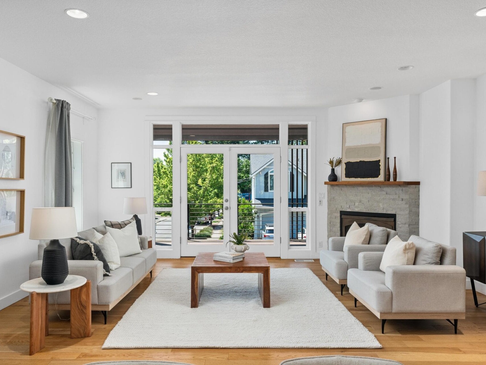 A bright, modern living room with white walls, light wood floors, two beige sofas, a wooden coffee table, a fireplace, large windows, and minimalist decor. Natural light fills the space.