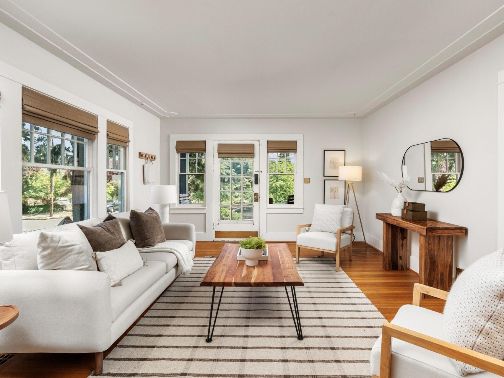 Bright living room with white sofas, wooden coffee table, striped rug, armchairs, and console table with decor. Large windows let in natural light and offer a view of greenery outside.