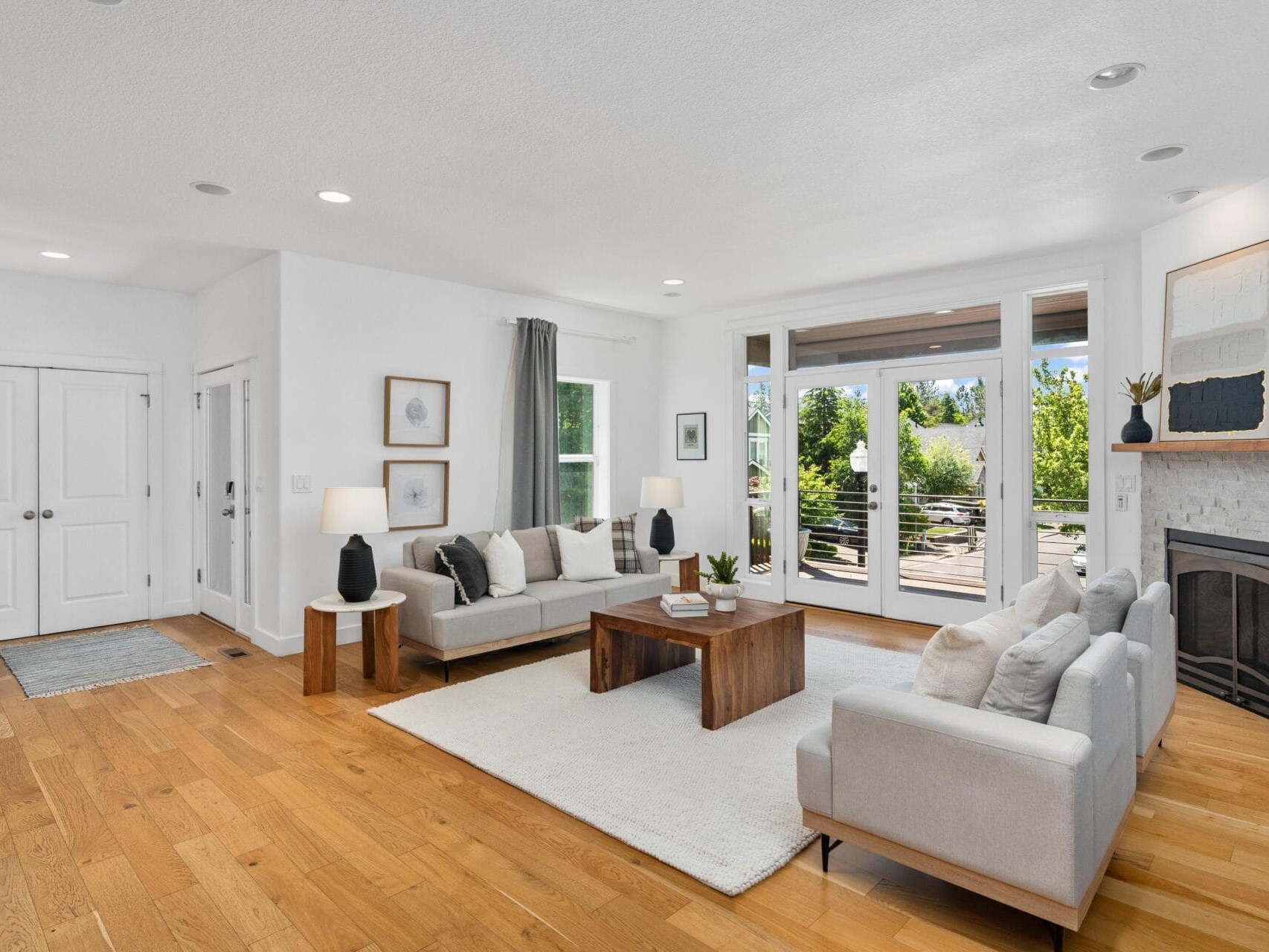 A modern living room with light wood floors, white walls, large windows, gray sofas, a wooden coffee table, lamps, and a fireplace with a stone surround and a mantel decorated with small plants and art.