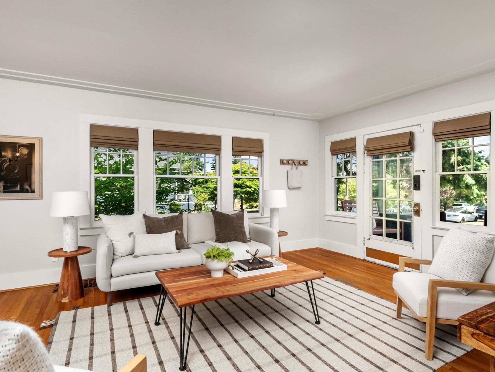 Bright living room with large windows, white walls, a white sofa with gray and brown pillows, a wooden coffee table, striped rug, armchair, side tables with lamps, and greenery visible outside.