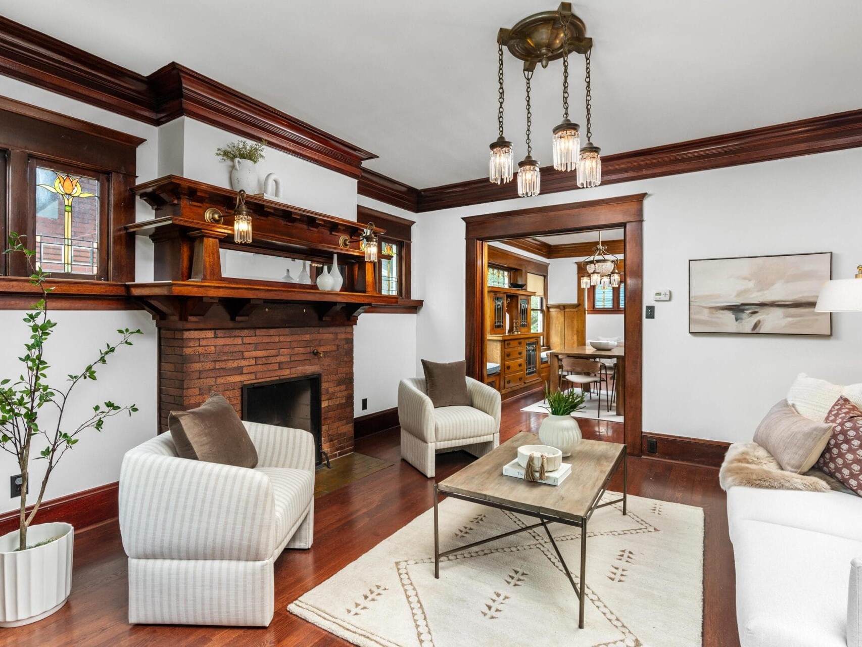 A cozy living room with dark wood trim, a brick fireplace with stained glass windows above, white armchairs, a white sofa, a coffee table, and a potted plant on hardwood floors.
