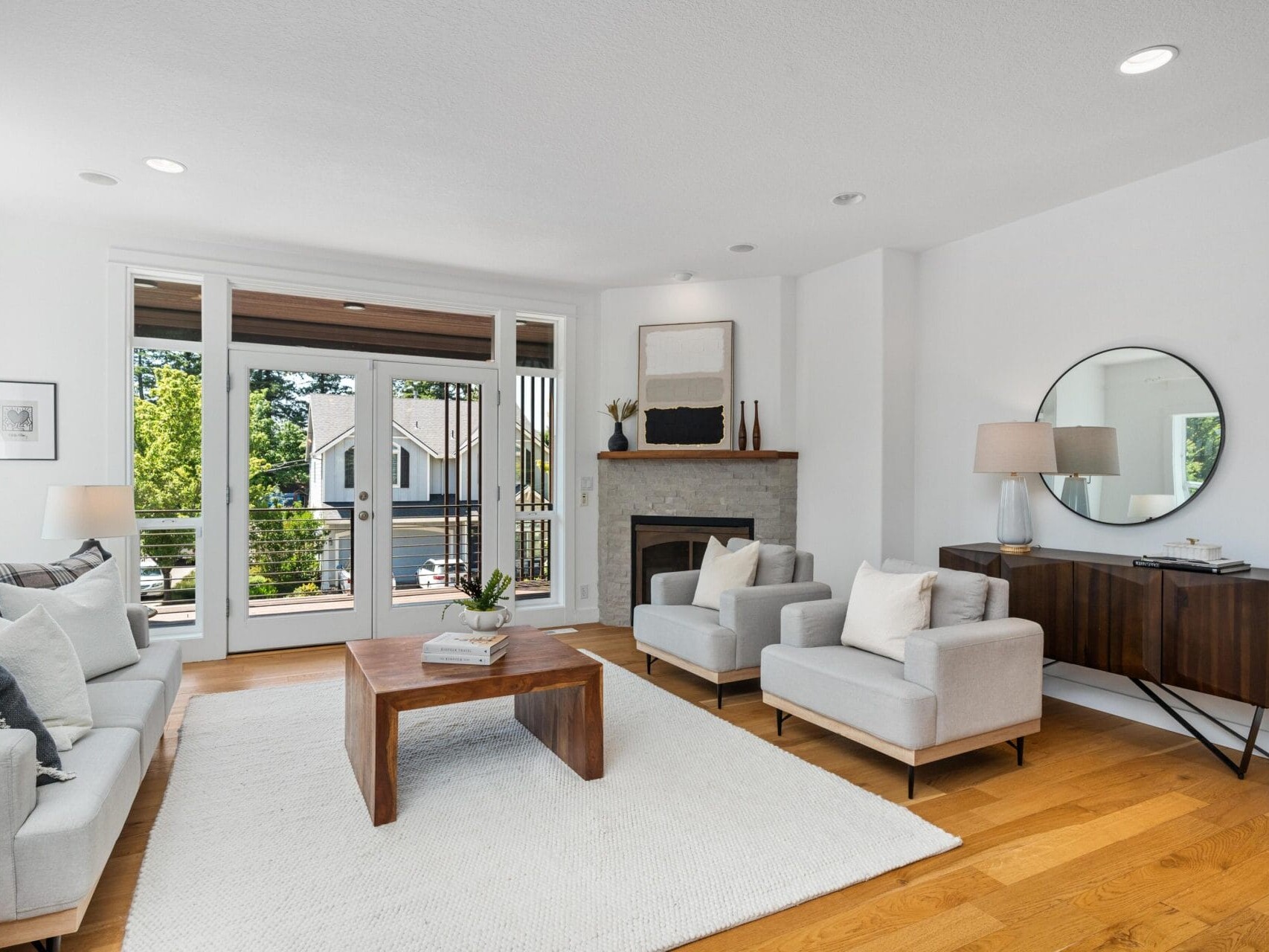A bright, modern living room with a white sofa, two gray armchairs, a wooden coffee table, a fireplace, a round mirror above a wooden console, and large windows letting in natural light.
