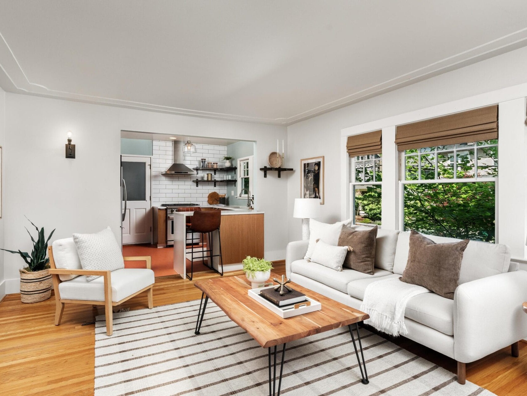 A bright, modern living room with a white sofa, wooden coffee table, armchair, striped rug, and large windows; the kitchen with white subway tiles and wood accents is visible in the background.