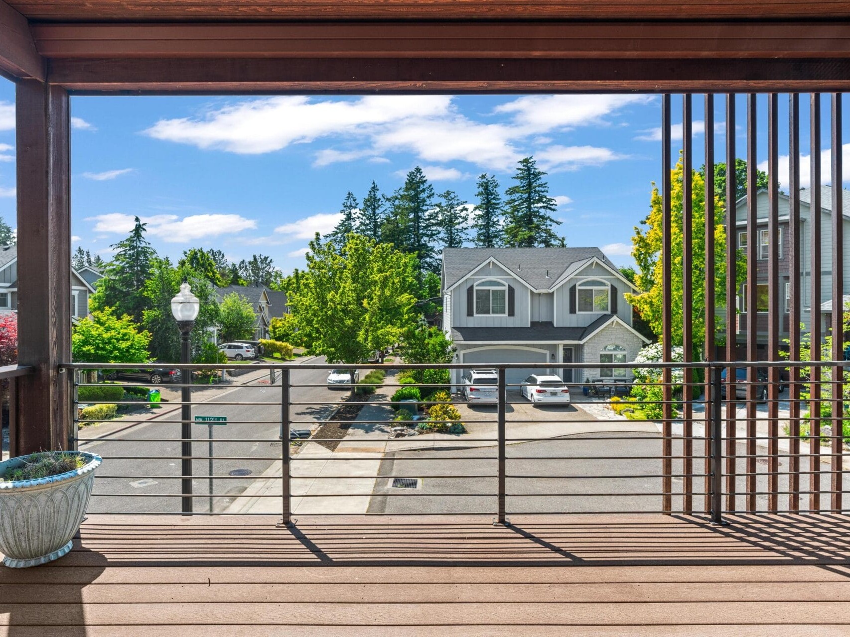 View from a balcony with wooden flooring and railing, overlooking a suburban street lined with trees, parked cars, and gray two-story houses under a bright, partly cloudy sky. A potted plant sits in the corner.