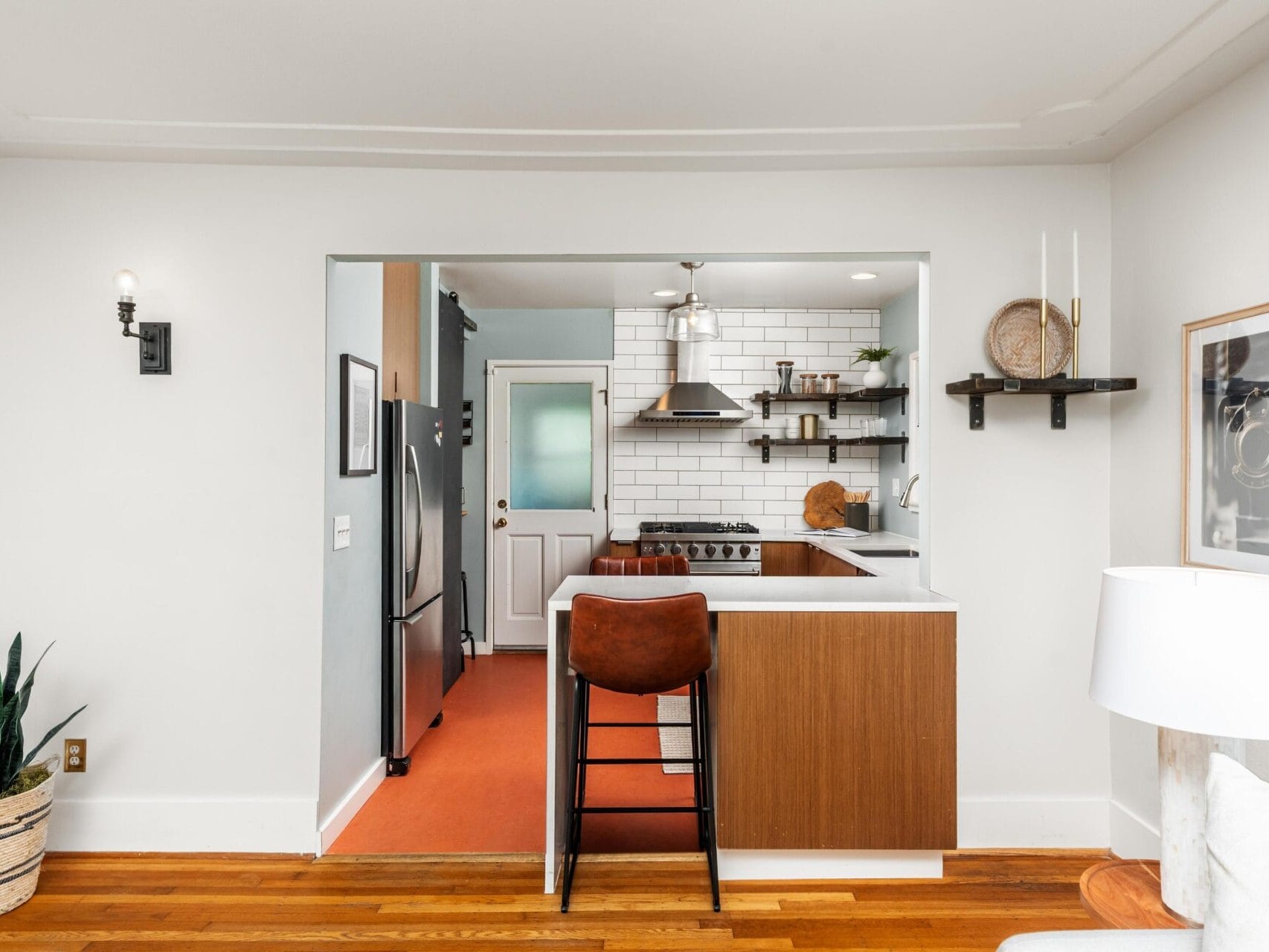A modern kitchen with white subway tile backsplash, stainless steel appliances, wooden cabinets, open shelves, and a brown bar stool at a white counter. Potted plants and wall art decorate the space.