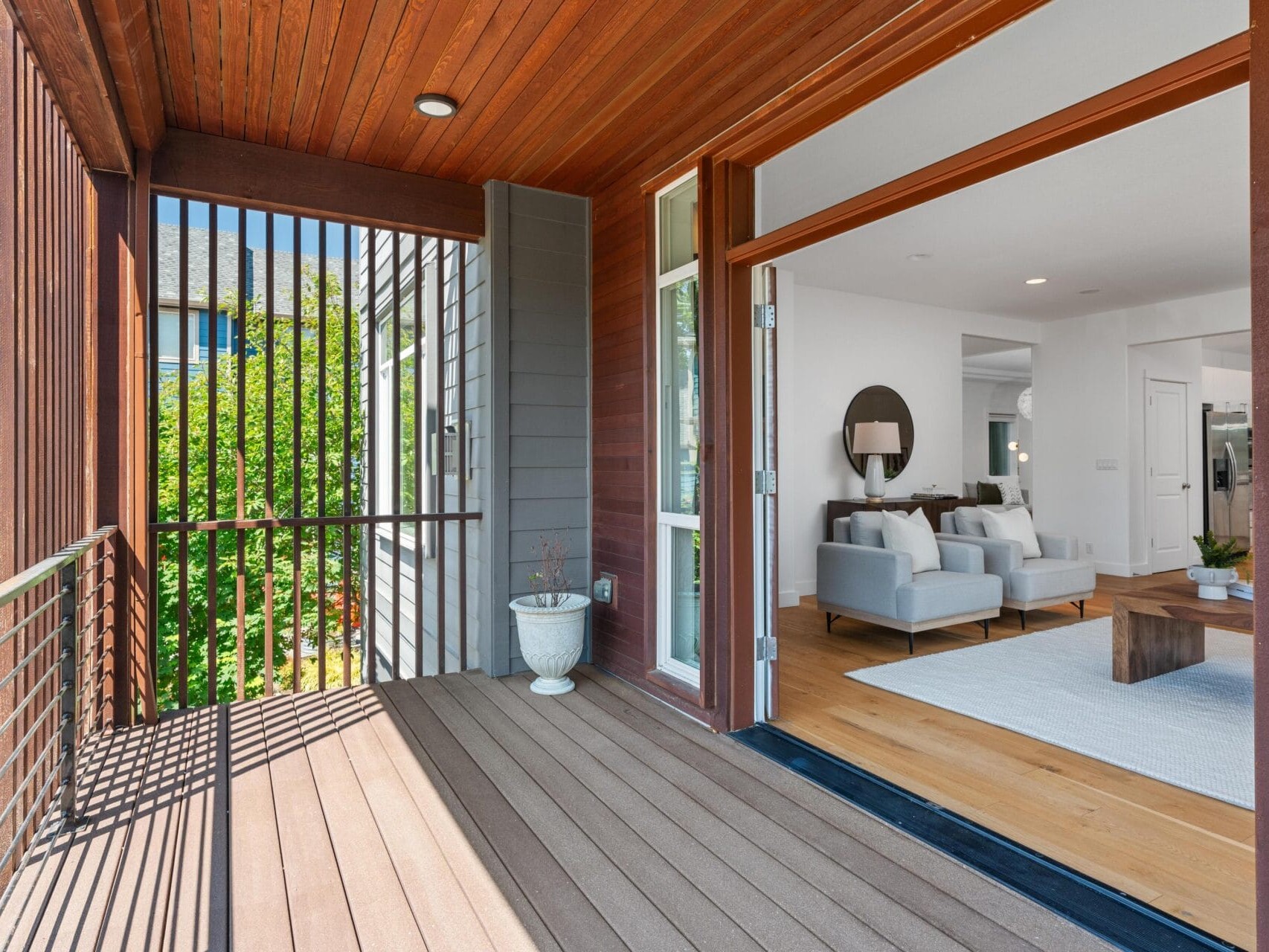 A modern home with an open sliding door leading from a covered wooden balcony into a bright living room with two light gray armchairs, a white rug, and minimalist decor.