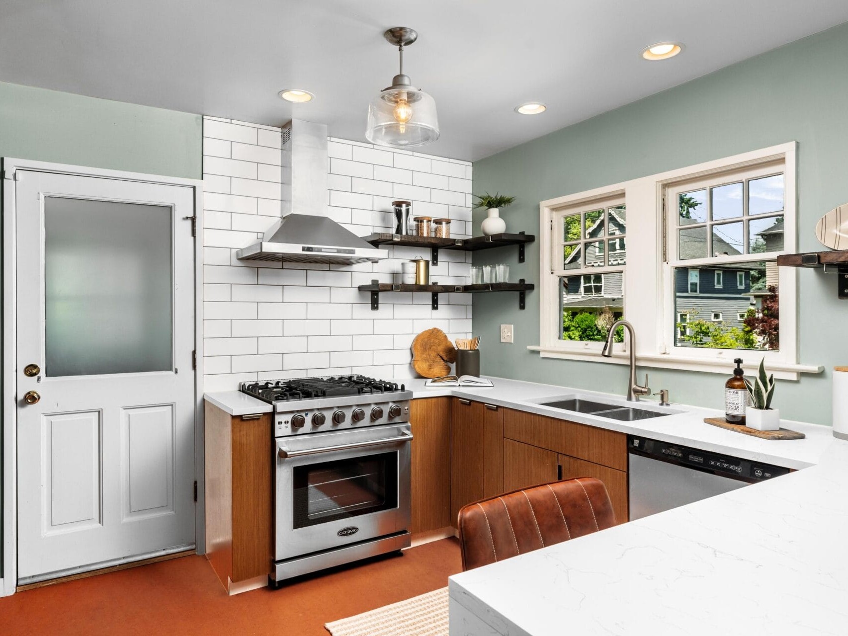 Modern kitchen with white subway tile backsplash, stainless steel stove and hood, open shelving, white countertops, a farmhouse sink, and large windows letting in natural light. A door leads to the outside.