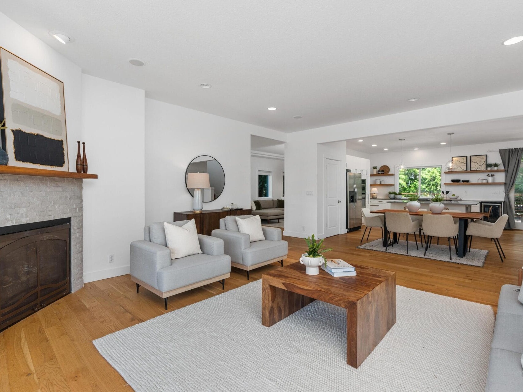 Modern open-concept living and dining area with light walls, wood flooring, a stone fireplace, neutral furniture, a wooden coffee table, and large windows letting in natural light.