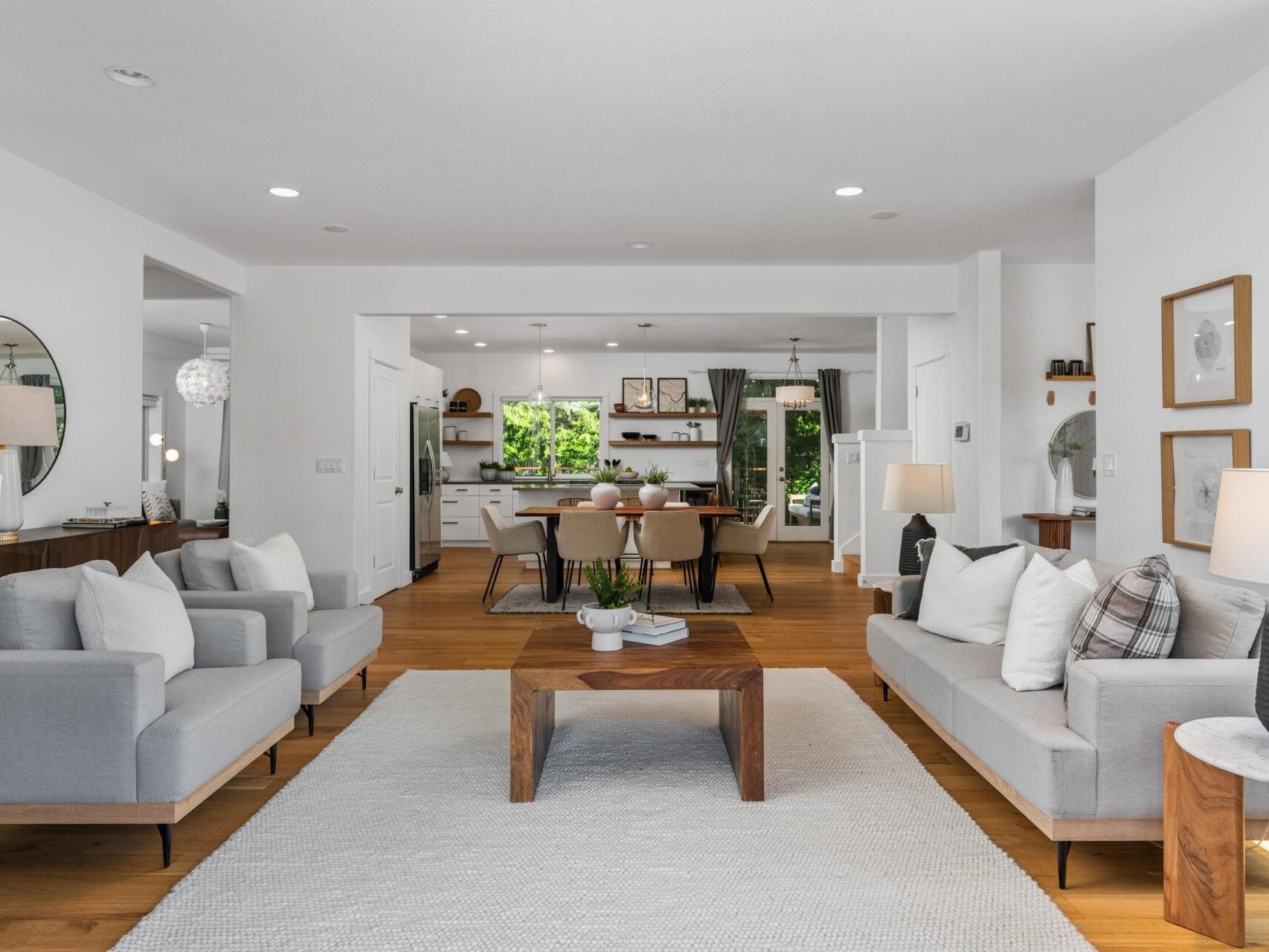 Modern living room with light gray sofas, wooden coffee table, and neutral decor. Open plan leads to a dining area with a wooden table and chairs, large windows, and minimalist accents. Light wood flooring throughout.