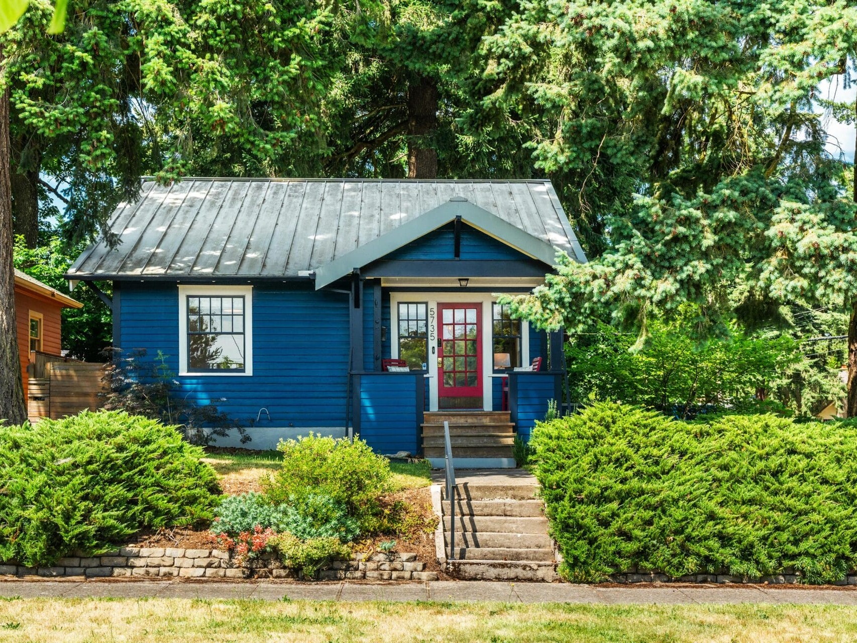 A small blue house with a metal roof and red front door sits among tall trees and lush green bushes, with a stone walkway and steps leading to the entrance.