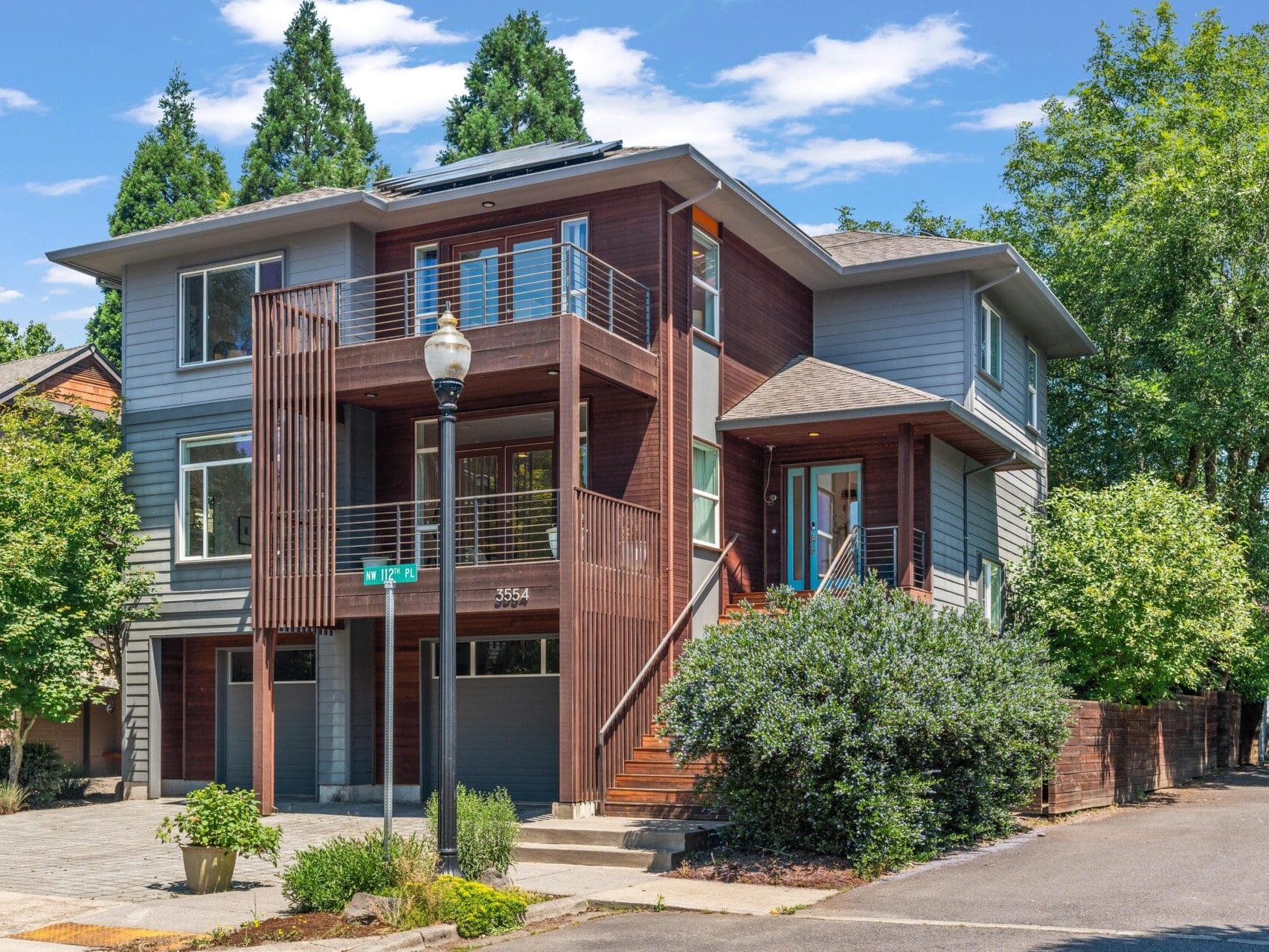 Modern two-story house with gray and wood siding, large windows, and a balcony. A streetlamp and green trees surround the property under a bright blue sky with scattered clouds.